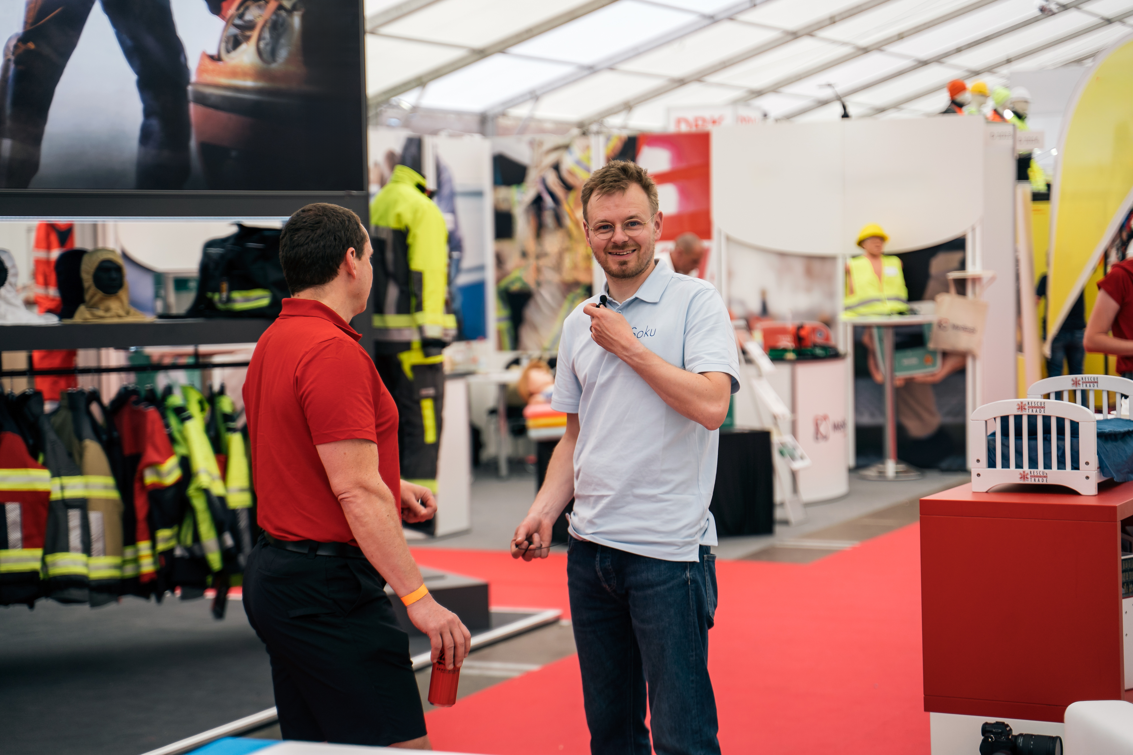 Two men interacting at a trade show or exhibition with various booths and displays in the background.